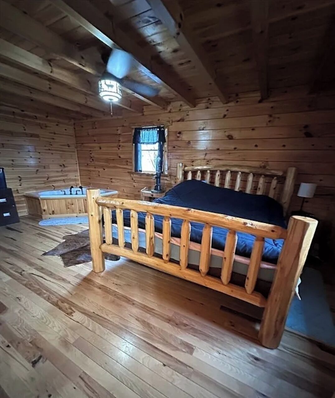 A wooden cabin room with a bed, a window, a ceiling fan, and a small table.