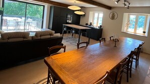 Un comedor amplio con una mesa y sillas de madera, una cocina al fondo y ventanas grandes que ofrecen luz natural.