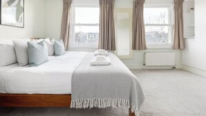 A hotel room with a large bed, two windows with curtains, a radiator, and a framed picture on the wall. - Stylish Flat in Wandsworth (London)
