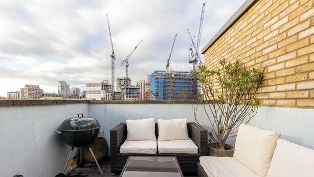 A rooftop patio with a grill, wicker sofa, and a view of the city skyline and construction cranes. - Stylish Flat in Wandsworth (London)