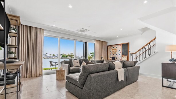 A spacious living room with a large grey sofa, a dining table, and a staircase leading to an upper floor.