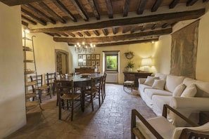 Un interno rustico con travi a vista in legno, pavimento in pietra, zona pranzo con tavolo e sedie, divano e mensola a parete.