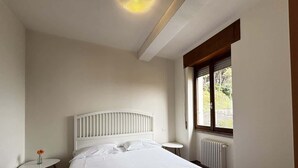 Un dormitorio con cama, una mesita de noche, una pequeña mesa con un florero, una ventana con vistas y un radiador.