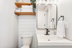 Een badkamer met een wit toilet, een witte wastafel, een spiegel en een houten plank met handdoeken en toiletartikelen.