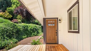 Una terraza de madera que conduce a una casa blanca con una puerta marrón y una ventana con persianas.