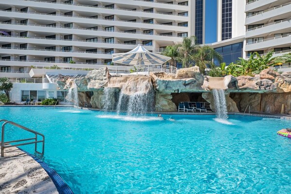 Piscina de hotel con caída de agua, rodeada por un edificio de varios pisos con balcones.