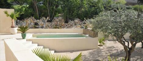 A pool area with steps leading down to a rectangular pool, surrounded by a stone wall and various plants.