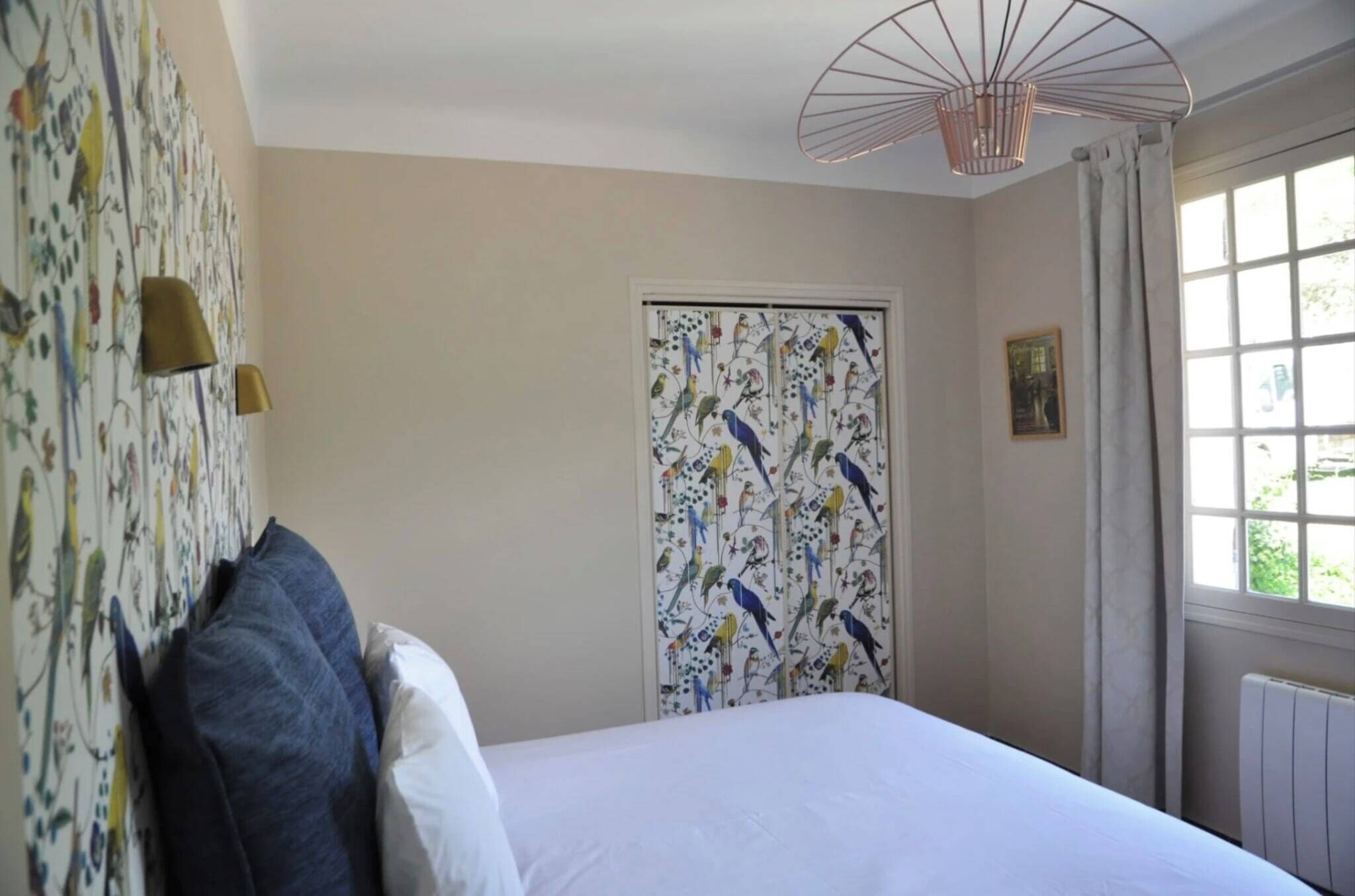 Un dormitorio con cama, ventana con cortinas, pared con diseño de pájaros y lámpara en el techo.
