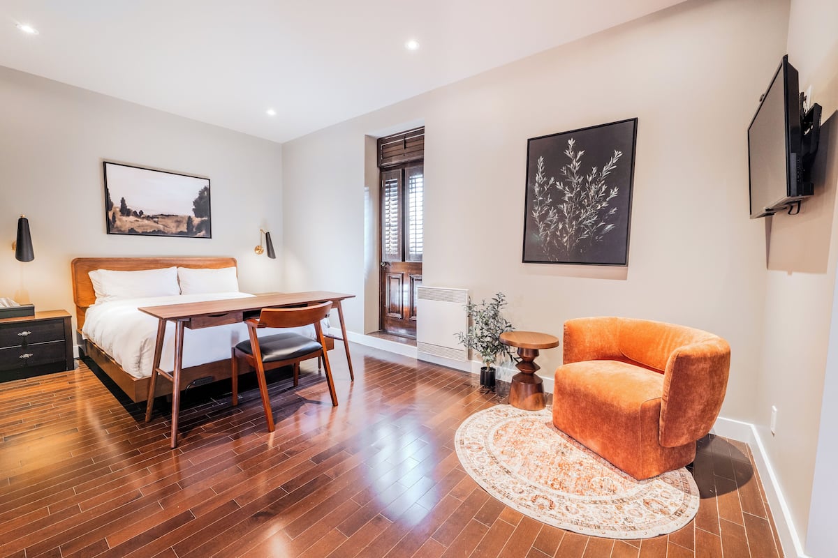 Une chambre d’hôtel moderne avec un plancher en bois, un lit recouvert de draps blancs, un bureau en bois avec une chaise, un fauteuil orange et un tapis rond.