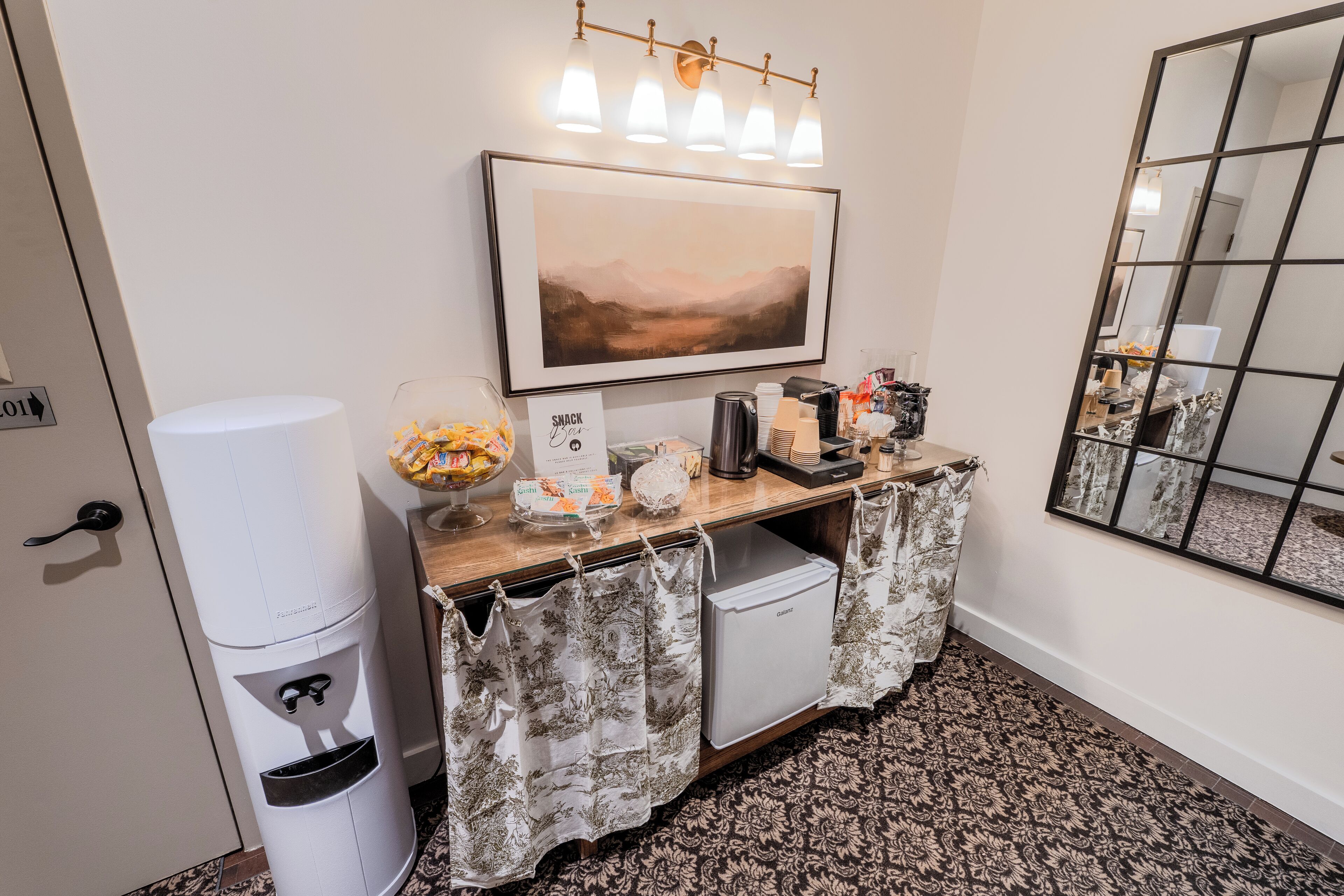 A hotel reception area with a framed landscape painting, a snack bar, a coffee station, and a trash bin.