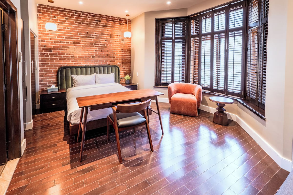 Une chambre avec un mur en briques, un lit, une table en bois, une chaise, une petite table ronde et une fenêtre avec des stores.