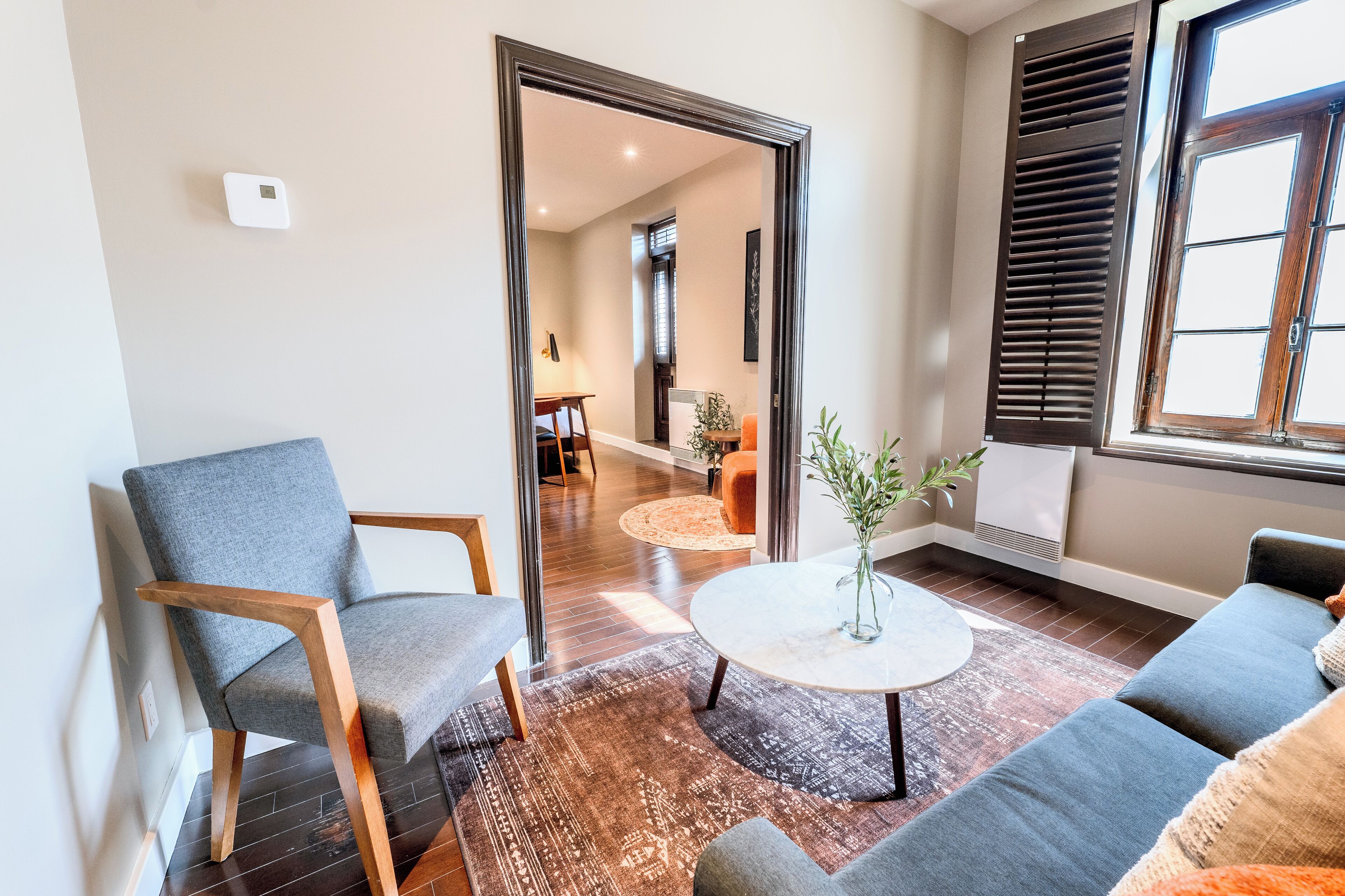 A modern living room with a grey sofa, a wooden chair, a round coffee table with a vase, and a large window with blinds.