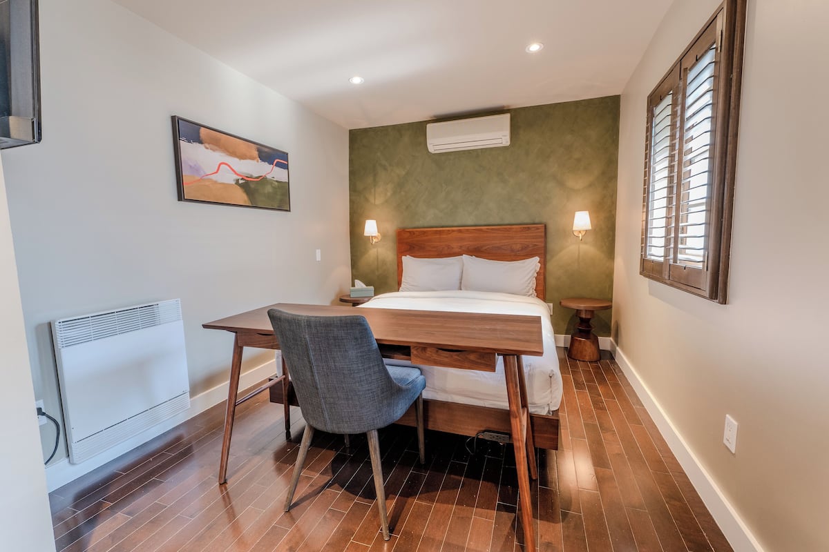 Une chambre à coucher comprenant un lit, un bureau, une chaise, un climatiseur, une lampe fixée au mur et une fenêtre avec des stores.