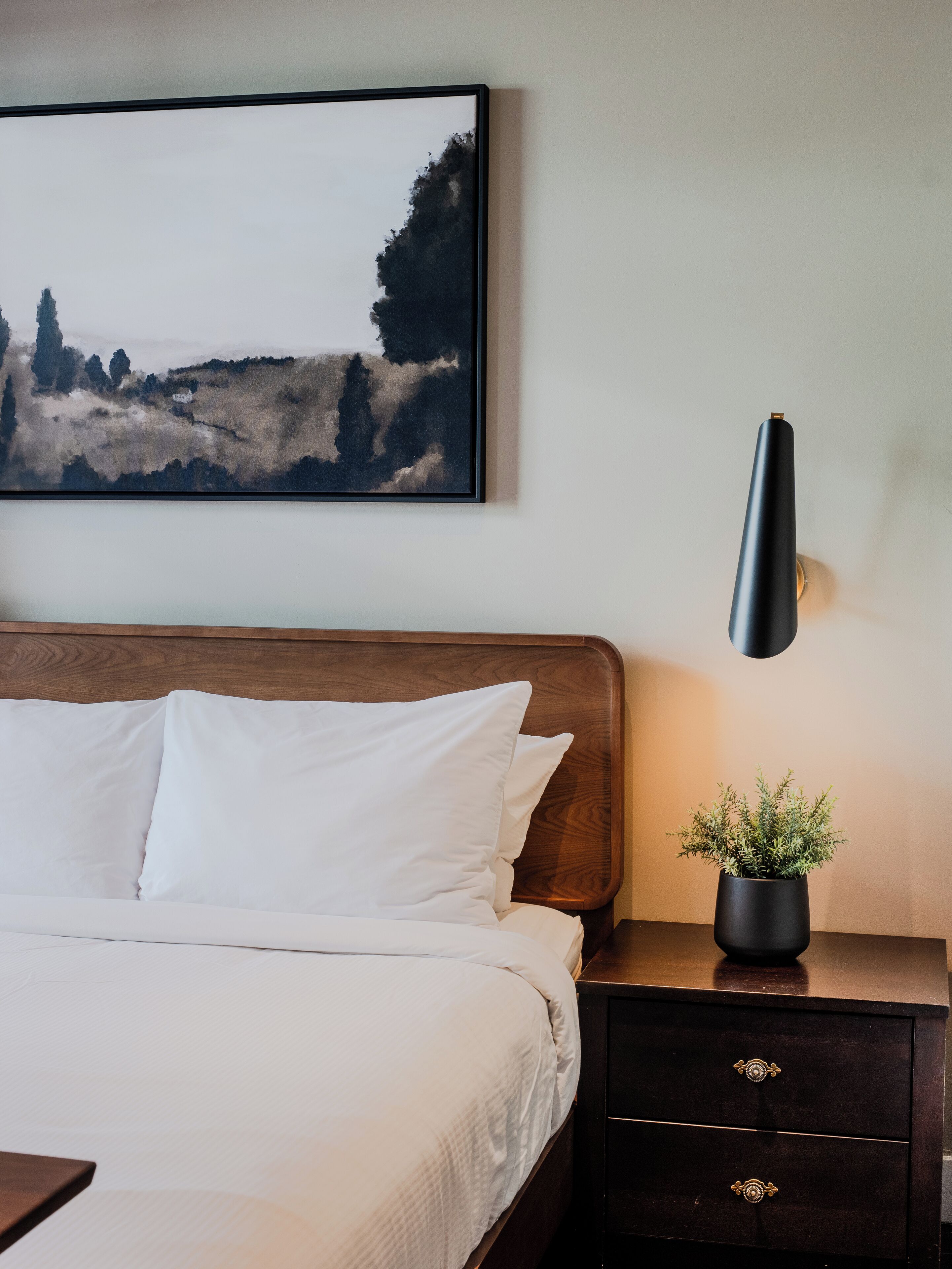 Un lit bien fait, avec du linge de lit blanc, une tête de lit en bois foncé et un tableau accroché au mur représentant un paysage.