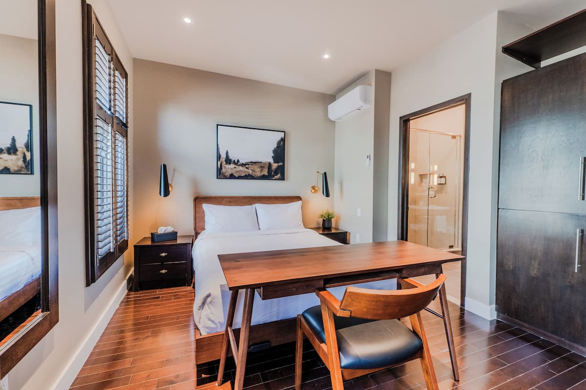 Une chambre d’hôtel comprenant un lit, une table en bois, une chaise, une table de chevet, un climatiseur fixé au mur et une porte donnant accès à une salle de bain.