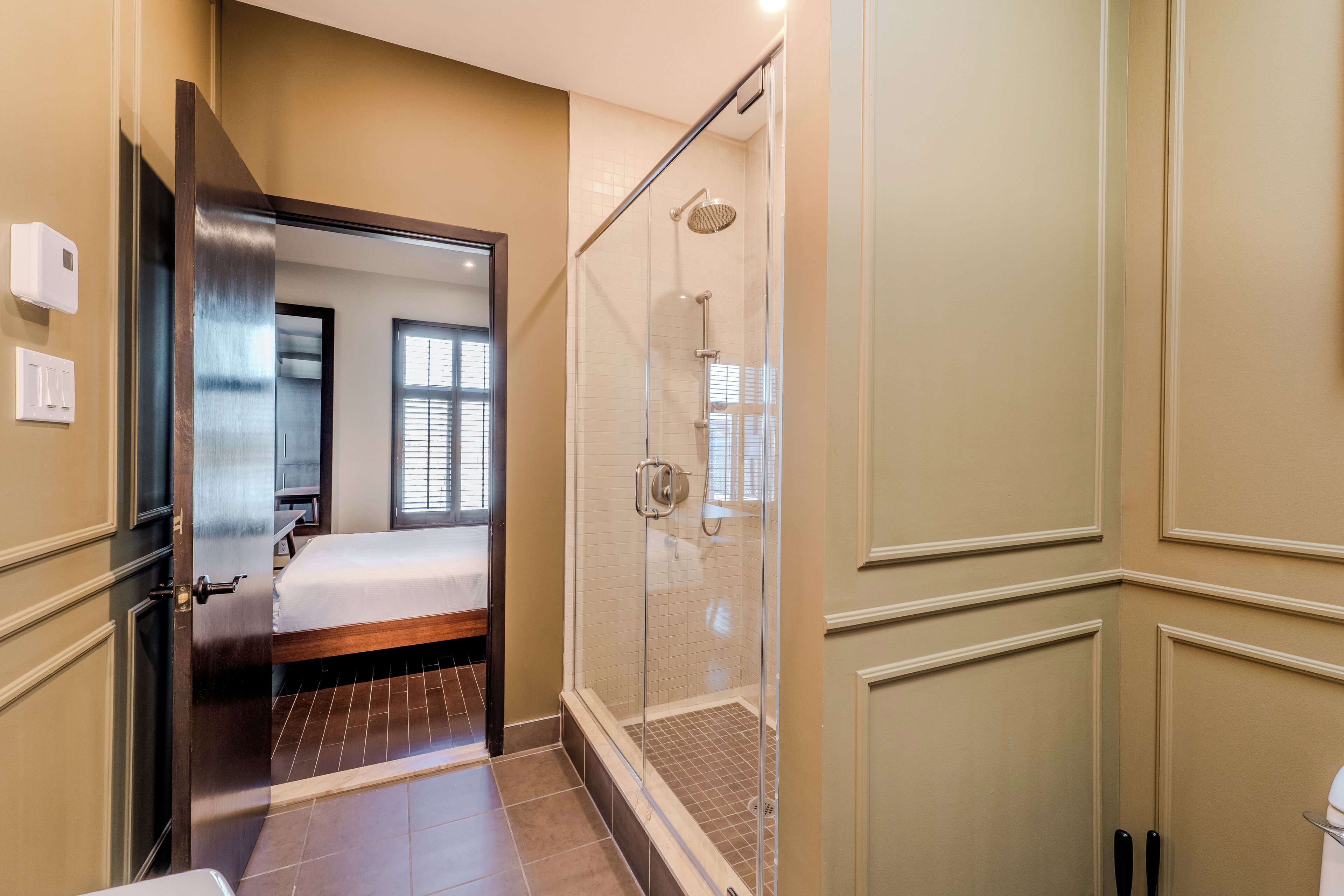 A modern bathroom with a glass-enclosed shower, a visible bed, and a door leading to another room.