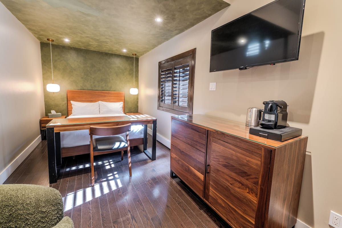 Une chambre d’hôtel comprenant un lit, un bureau, une chaise, une commode en bois, un téléviseur et une fenêtre avec des stores.
