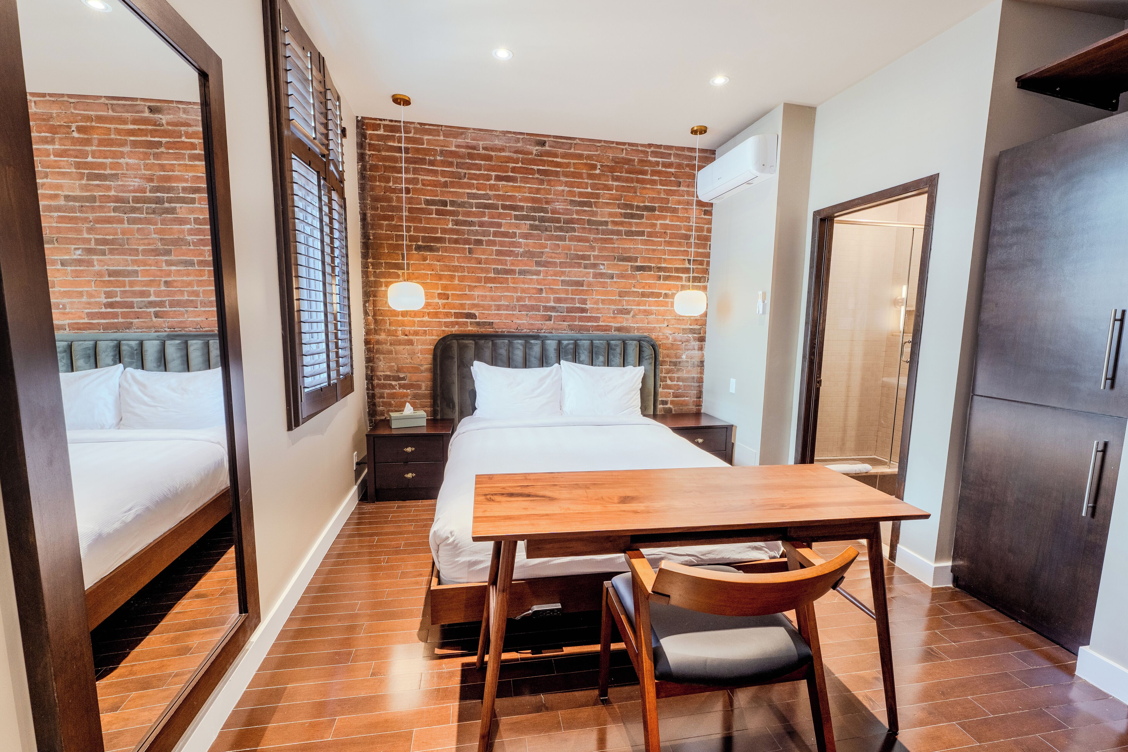 Habitación de hotel con pared de ladrillo, cama con ropa blanca, mesa de madera con silla y un armario.