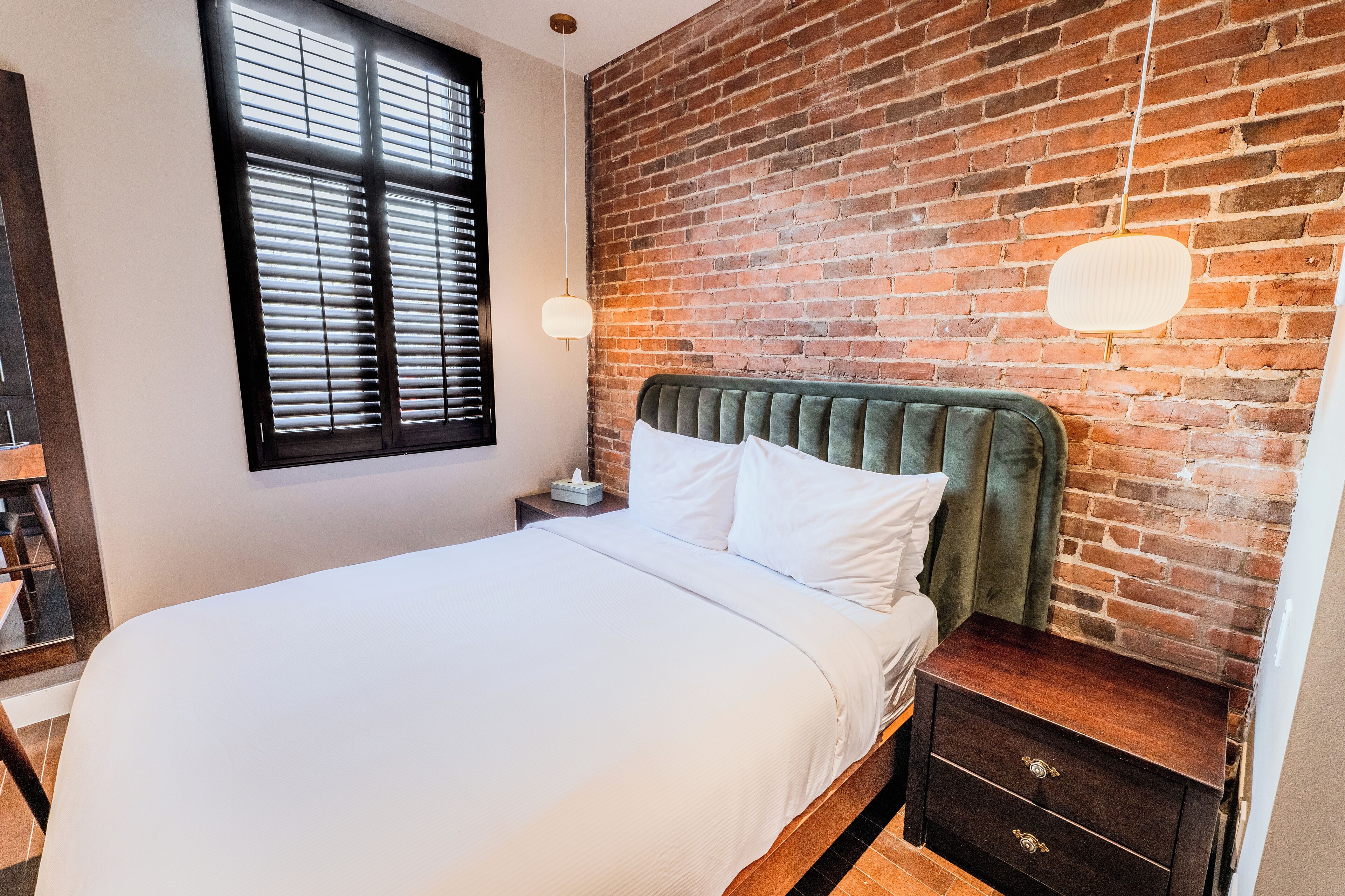 A neatly made bed with white linens, a brick wall, and a nightstand with a lamp.