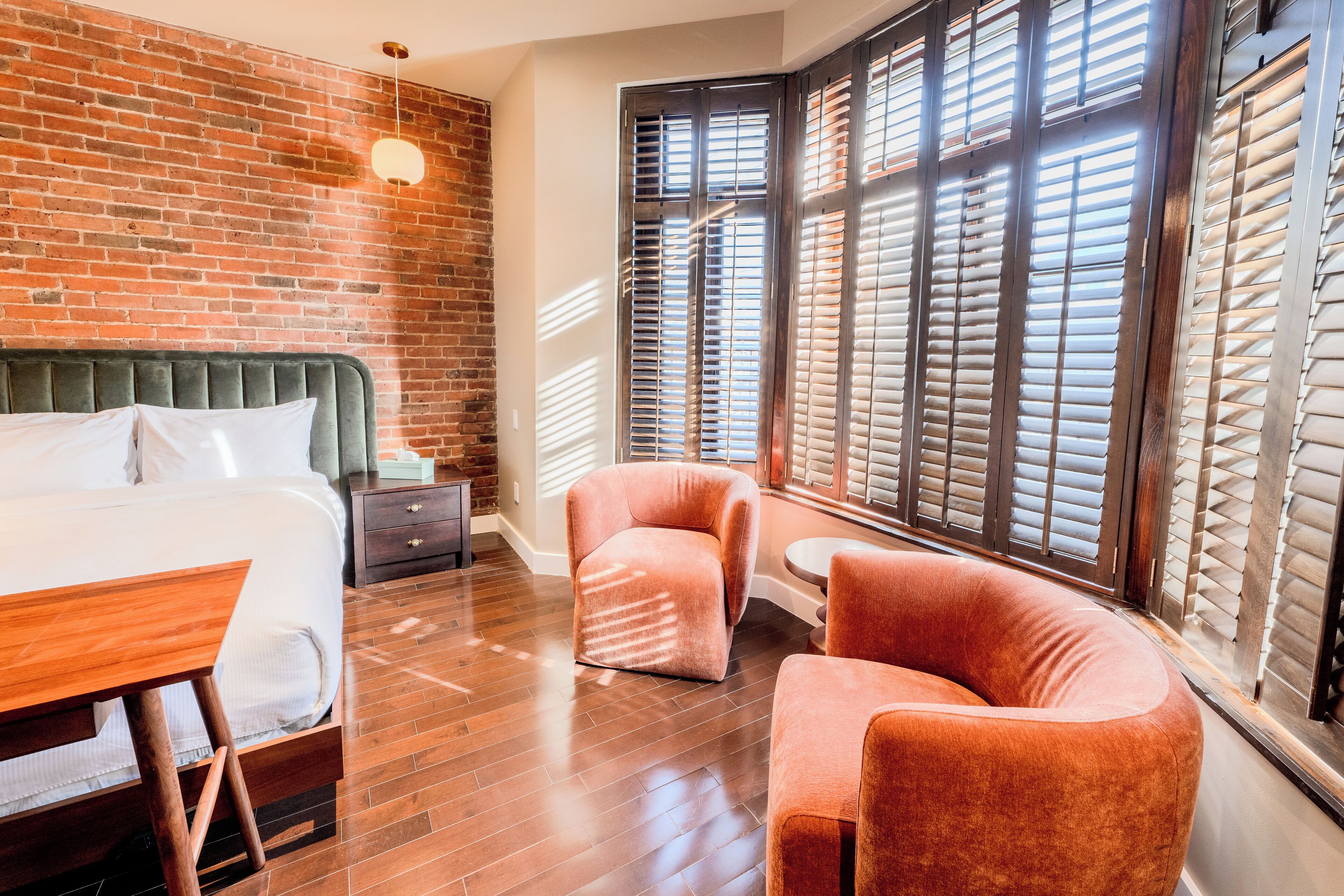 Une chambre d’hôtel avec un mur en briques, un grand lit, un bureau en bois, deux fauteuils et une fenêtre avec des stores.