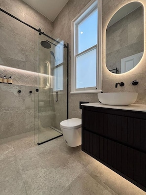 Un baño moderno con ducha envuelta en vidrio, inodoro blanco, tocador de madera oscura y un espejo grande con marco iluminado.