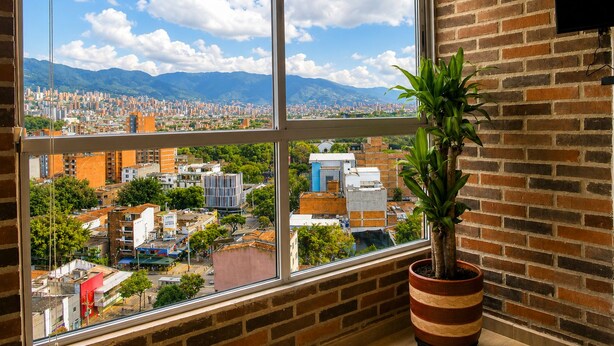 Habitación con un amplio ventanal que ofrece vista panorámica de la ciudad, una planta en maceta y una pared de ladrillo.