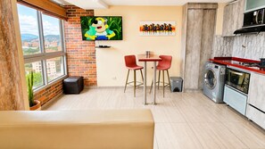A modern living room with a large window, a flat-screen TV, a dining area with red chairs, a sofa, and a kitchen with a washing machine and microwave.