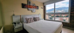 A hotel room with a bed, a nightstand, a phone, and a window with a city view. - Laureles elegant Spring City Treat  (Medellín)
