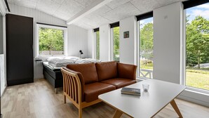 A modern bedroom with a bed, a brown leather sofa, a white table, and large windows offering a view of greenery. - Beautiful Home In Ebeltoft With Sauna (Ebeltoft)