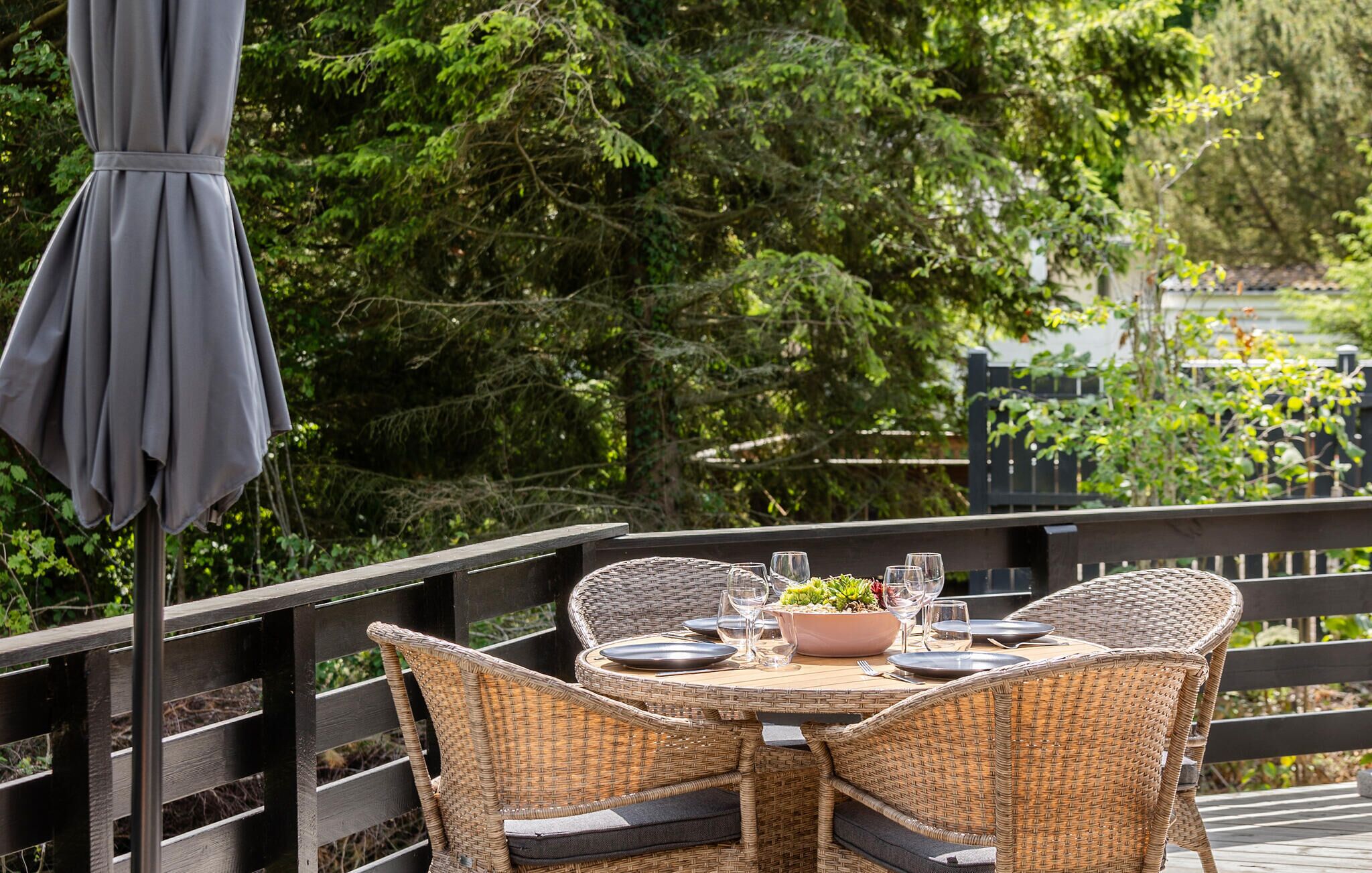 Een terras met rieten stoelen en een ronde tafel voor twee personen, omgeven door groen.