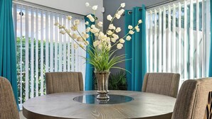 A round table with a glass top, surrounded by four upholstered chairs, in a room with teal curtains and a potted plant with white flowers. - 5 Bedroom Pool Home in Gated Community Near Disney (Kissimmee)