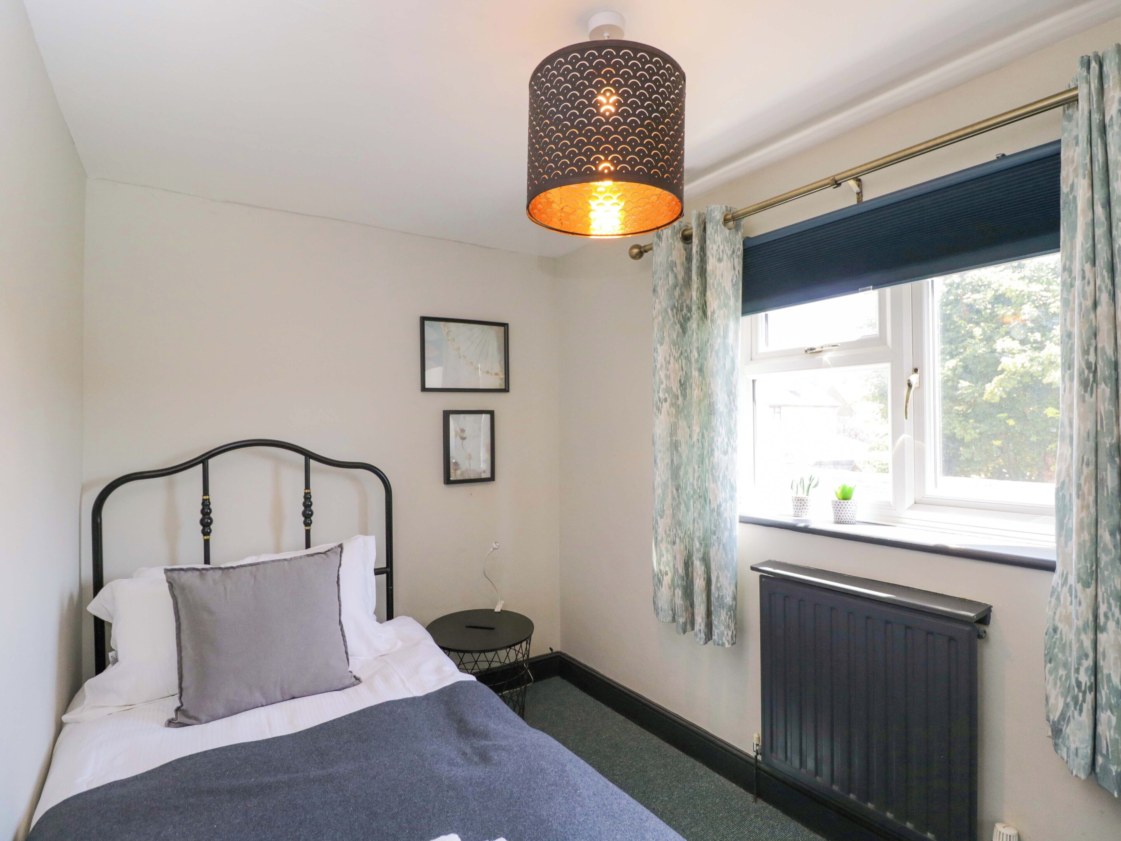 Une chambre à coucher comprenant un lit, une table de chevet, un radiateur, une fenêtre avec des rideaux et un luminaire suspendu.