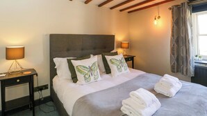 A hotel room with a large bed, two bedside tables with lamps, a window with curtains, and exposed wooden beams on the ceiling. - Elm Cottage (Moreton-in-Marsh)