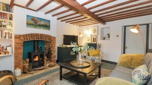 A cozy living room with exposed wooden beams, a brick fireplace, a sofa, and a coffee table with decorative items. - Elm Cottage (Moreton-in-Marsh)