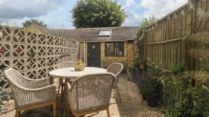 A patio with wicker furniture and a table, surrounded by a wooden fence and potted plants. - Elm Cottage (Moreton-in-Marsh)