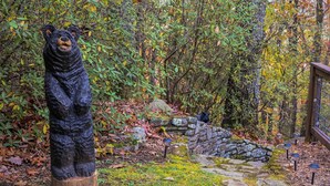 Una escultura de oso de madera en un área boscosa con muros de piedra y una valla de madera.