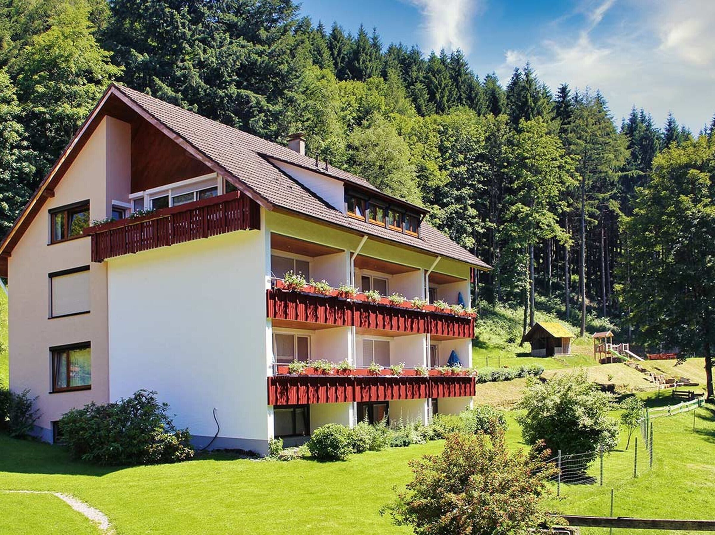 A two-story building with a balcony, surrounded by lush greenery and a forested backdrop.