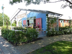 A small, single-story building with a 'Take Away' sign, a covered outdoor seating area, and a garden with various plants.