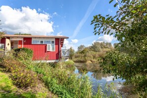 A red building with a window and a door, surrounded by greenery and a body of water.