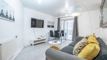 A modern living room with a grey sofa, a flat-screen TV, a dining area with chairs, and a large window.