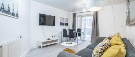 A modern living room with a grey sofa, a flat-screen TV, a dining area with chairs, and a large window.