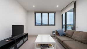 A modern living room with a flat-screen TV, a white coffee table, a brown sofa, and a large window offering a city view. - Convenient City Apartment in the Heart of Braddon (Braddon)