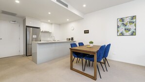 A modern kitchen with white cabinets, a stainless steel refrigerator, and a wooden dining table with blue chairs. - Convenient City Apartment in the Heart of Braddon (Braddon)