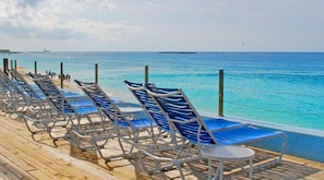 Une rangée de fauteuils bleus est disposée sur une terrasse en bois, offrant une vue imprenable sur une mer d’un bleu limpide.