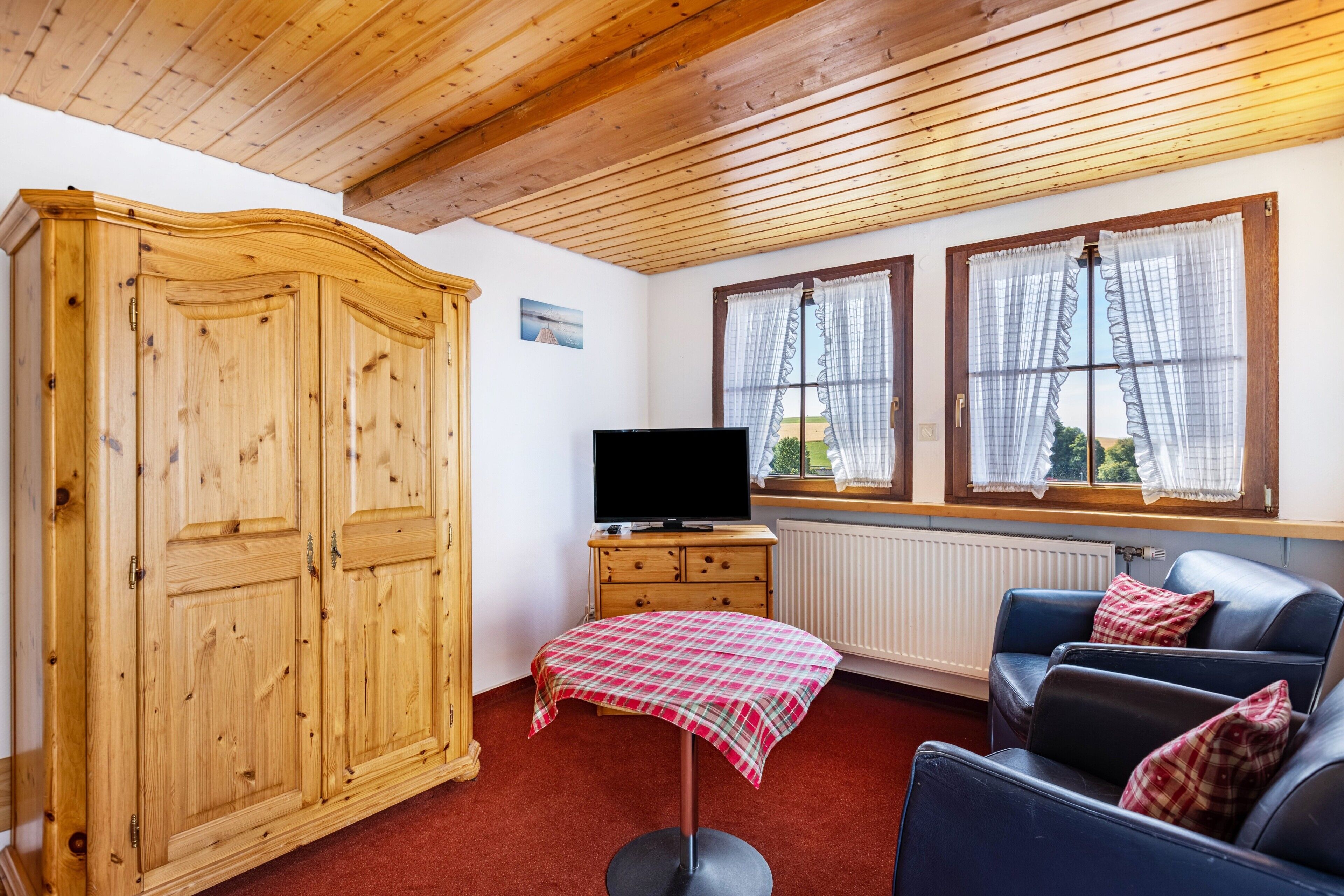 A room with a wooden wardrobe, a television on a wooden cabinet, a table with a plaid tablecloth, and a sofa with pillows.