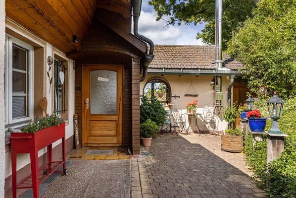 A wooden house with a tiled roof, a wooden door, and a small garden area with potted plants and a table.