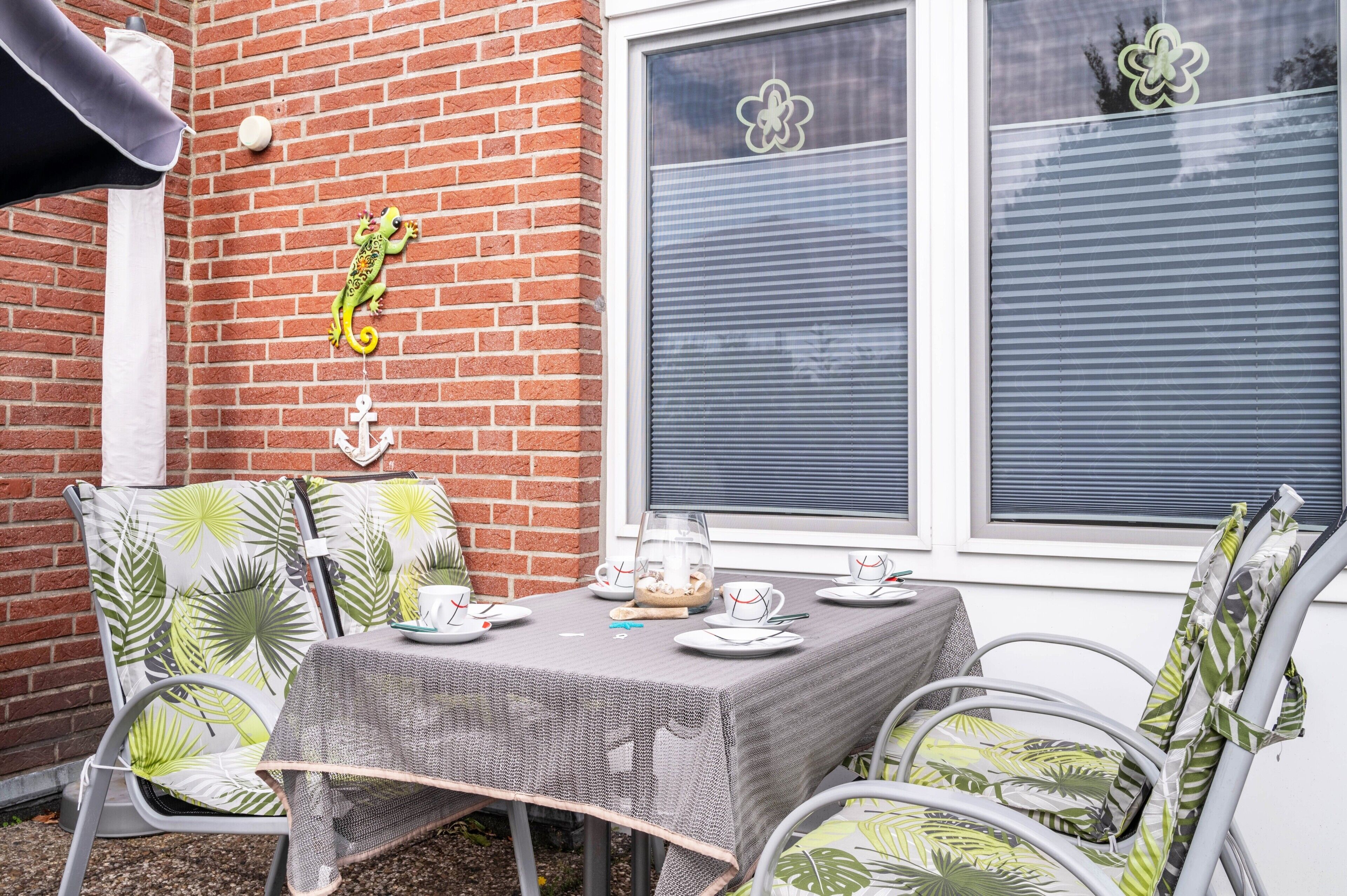 Eine Terrasse mit Tisch für zwei Personen, Stühle mit Blattmuster, einer Backsteinmauer und einem Fenster mit floralem Dekor.