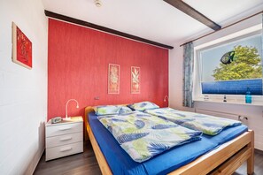 Ein Schlafzimmer mit roter Akzentwand, einem Bett mit blaugrüner Bettdecke mit Blattmuster, einem weißen Nachttisch mit Lampe und einem Fenster mit Blick auf Bäume.