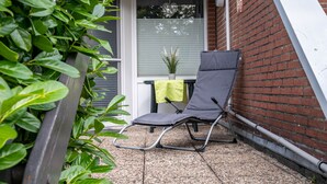 Een balkon met een grijze lounge stoel, een plant in pot en een raam met zonwering.
