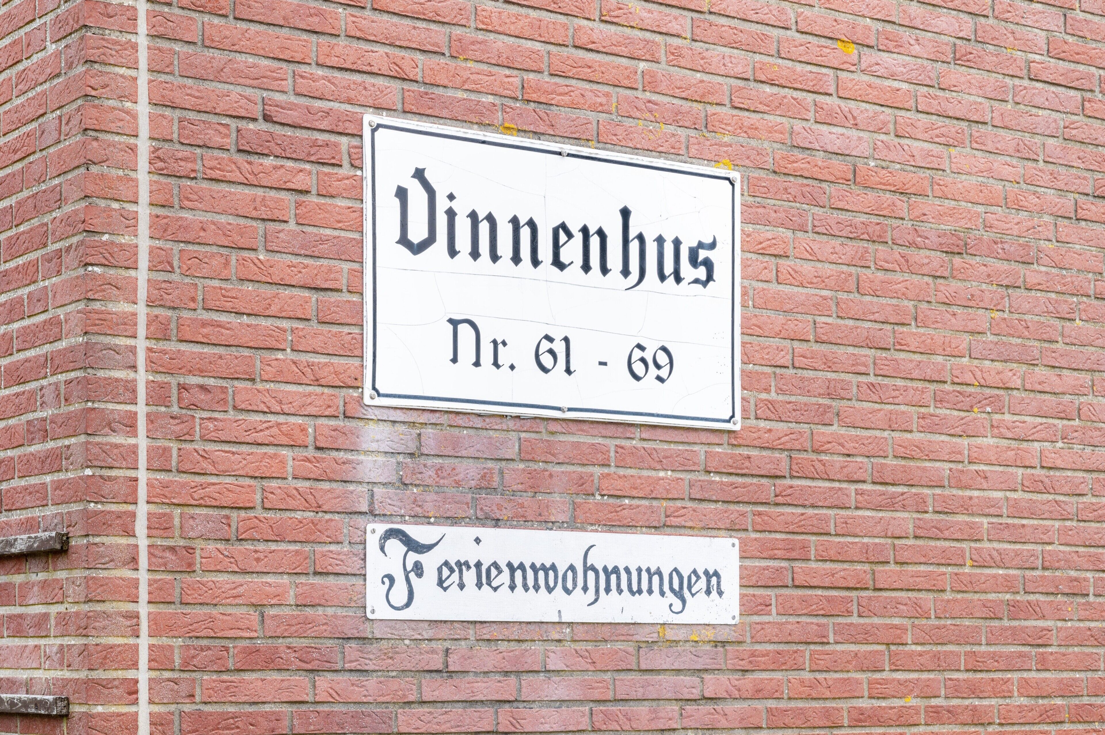 Ein Schild an einer Backsteinmauer mit dem Text 'Vinnenhus Nr. 61-69' und 'Ferienwohnungen', das auf ein Anwesen mit nummerierten Zimmern hinweist.